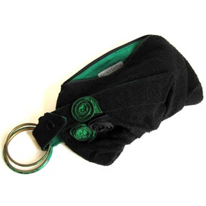 Puede incluir: Clutch de tela negra con forro verde y adorno de flores de tela verde. El clutch tiene un asa de anillo de metal dorado y una etiqueta que dice "Megan Leone".