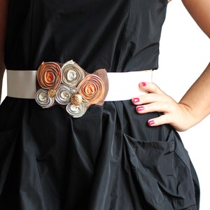 Puede incluir: Un cinturón blanco con una hebilla grande y decorativa hecha de materiales reutilizados, incluyendo cremalleras y detalles metálicos. La hebilla es un conjunto de círculos naranja, marrón y plateado y se lleva sobre un vestido negro.