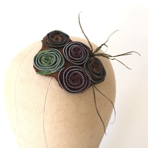 May include: A brown fabric base with five fabric flower embellishments made from zippers. The flowers are in shades of green, purple, and brown. A single feather is attached to the base.