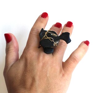 May include: A black ring with a large, irregular, gray stone wrapped in gold wire. The ring is worn on a finger with red nail polish.