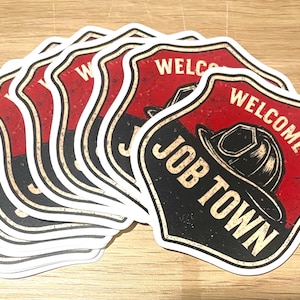 May include: A stack of shield-shaped stickers with a red and black design. The stickers feature the text "WELCOME TO JOB TOWN" and an illustration of a firefighter's helmet. The stickers have a distressed, vintage look.