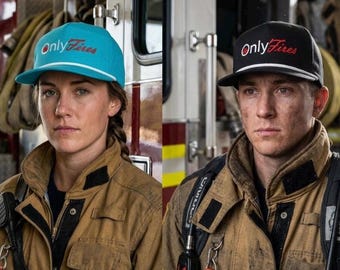 May include: Two people wearing tan firefighter jackets and caps with the text "Only Fires". One cap is teal with white trim, the other is black with white trim. Both are looking at the camera.