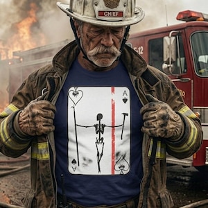 Peut inclure: Un pompier portant un casque et une veste, avec un t-shirt bleu marine orné d'un motif de carte à jouer. La carte représente un squelette avec une bande rouge et l'As de Pique et de Coeur. Un camion de pompiers est en arrière-plan.