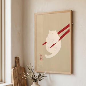 Könnte beinhalten: Ein gerahmter Kunstdruck mit einer minimalistischen Illustration einer runden, cremefarbenen Katze, die von zwei roten Essstäbchen vor einem hellbeigen Hintergrund gehalten wird. Die chinesischen Schriftzeichen 饺子 sind unten links rot aufgedruckt.