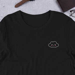 Könnte beinhalten: Schwarzes T-Shirt mit einem kleinen gestickten Wolken-Design mit einem lächelnden Gesicht. Das Shirt besteht aus einem weichen Material und hat einen klassischen Rundhalsausschnitt. Das Shirt wird auf einer neutralen Oberfläche mit anderem Zubehör präsentiert.
