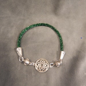 K&ouml;nnte beinhalten: Ein gr&uuml;nes geflochtenes Armband mit silbernen Endkappen und einem silbernen Anh&auml;nger in der Mitte. Der Anh&auml;nger ist ein kreisf&ouml;rmiges Design mit einem Mandala-Muster.