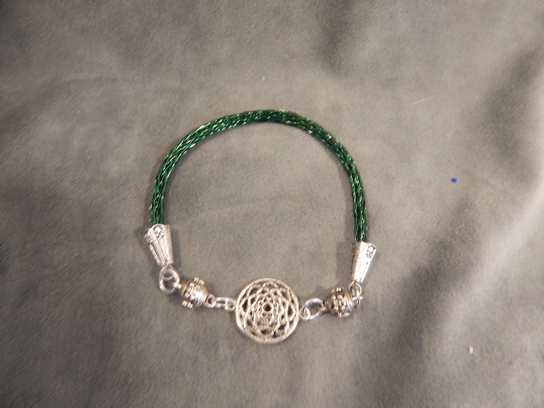 K&ouml;nnte beinhalten: Ein gr&uuml;nes geflochtenes Armband mit silbernen Endkappen und einem silbernen Anh&auml;nger in der Mitte. Der Anh&auml;nger ist ein kreisf&ouml;rmiges Design mit einem Mandala-Muster.