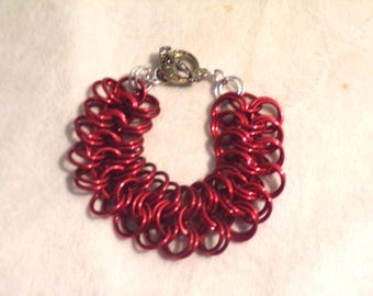Könnte beinhalten: Ein rotes Kettenarmband mit einem silbernen Verschluss. Das Armband besteht aus kleinen, ineinandergreifenden roten Metallringen. Der Verschluss ist silberfarben mit einem dekorativen, verzierten Design. Es ist ein Schmuckstück.