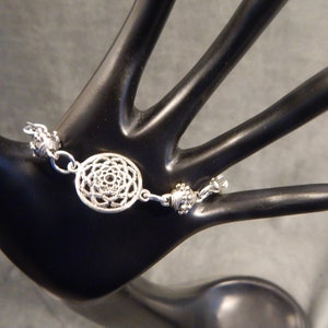 K&ouml;nnte beinhalten: Silberarmband mit einem runden Anh&auml;nger mit Mandala-Design. Der Anh&auml;nger ist durch eine Kette mit kleinen silbernen Perlen mit dem Armband verbunden.