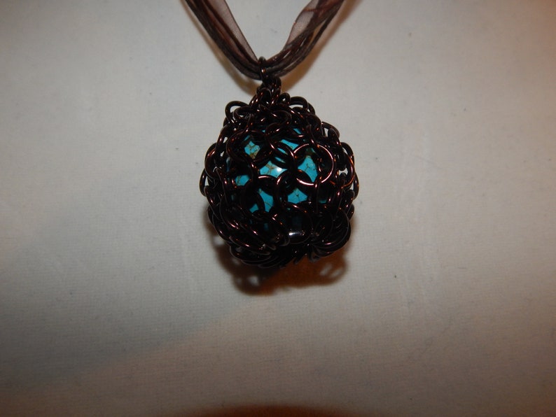 May include: A turquoise gemstone pendant encased in a brown wire cage, hanging from a brown ribbon.