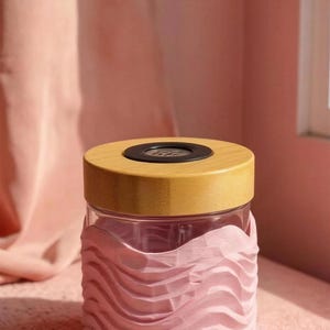 Puede incluir: Un tarro de cristal transparente con tapa de madera y un diseño rosa en forma de ola alrededor de la base. La tapa tiene un centro negro. El tarro está sobre una superficie rosa, con una cortina rosa al fondo.