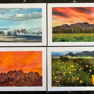 May include: Four framed photographs of a mountain range with different sky and landscape scenes. The top left photo shows a mountain range with clouds and a blue sky. The top right photo shows a mountain range with a red sunset sky. The bottom left photo shows a mountain range with an orange sunset sky. The bottom right photo shows a mountain range with a field of yellow flowers in the foreground.