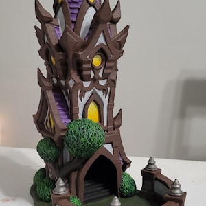 May include: A detailed, miniature haunted house model. The structure features a brown exterior with purple roofing and yellow-lit windows. The model includes a small yard with green shrubbery and a decorative fence.
