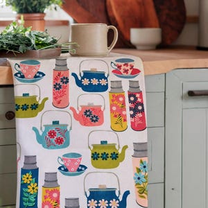 May include: A white kitchen towel featuring a repeating pattern of colorful teapots, teacups on saucers, and thermal flasks. The illustrations include various floral motifs in shades of blue, pink, yellow, and green, arranged across the fabric surface.