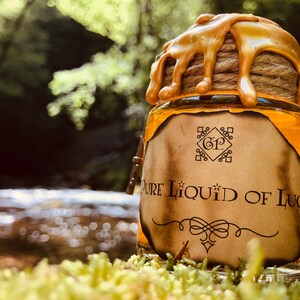 Könnte beinhalten: Ein Glasgefäß mit einem goldenen Deckel und einem Etikett mit der Aufschrift "Pure Liquid of Luck". Das Gefäß steht auf einem Moosbett in einer Waldlandschaft.