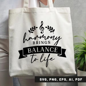 May include: Cream-colored tote bag with the text "harmony brings balance to life" in black script and a musical note and leaf design. The bag is held by a person and has a simple, minimalist design.
