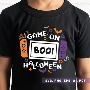 May include: Black t-shirt with a Halloween-themed graphic. The design includes the words "GAME ON BOO! HALLOWEEN" with game controller illustrations, bats, and spiderwebs. The shirt is made of a soft material.