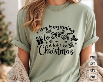 May include: Sage green t-shirt with the text "It's beginning to COST a lot like Christmas" in black. The design includes a gift box, snowflakes, and holly. The shirt has rolled-up sleeves.