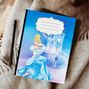 Puede incluir: Un cuaderno azul con Cenicienta sosteniendo un perro blanco y un castillo de fondo. La portada tiene una etiqueta blanca con l&iacute;neas para un nombre. Un bol&iacute;grafo negro descansa sobre una manta gris.