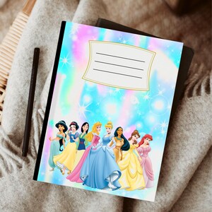 Puede incluir: Cuaderno con una colorida cubierta hologr&aacute;fica con ilustraciones de varias princesas con sus ic&oacute;nicos vestidos. El cuaderno tiene un &aacute;rea de etiqueta con l&iacute;neas y un lomo negro. Un bol&iacute;grafo negro descansa junto al cuaderno.