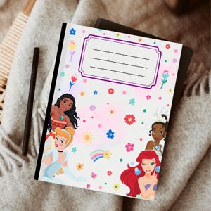 Puede incluir: Un cuaderno con ilustraciones de princesas animadas, incluyendo Moana, Cenicienta, Ariel y Tiana. La portada es blanca con un fondo rosa y acentos florales y arco&iacute;ris coloridos. Un bol&iacute;grafo negro descansa junto al cuaderno.