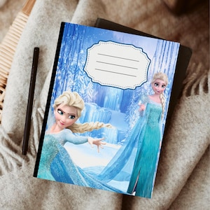 Puede incluir: Un cuaderno con una ilustraci&oacute;n azul y blanca de un personaje de la pel&iacute;cula Frozen. La portada tiene un &aacute;rea de etiqueta con l&iacute;neas para escribir. Un bol&iacute;grafo negro descansa sobre la manta.