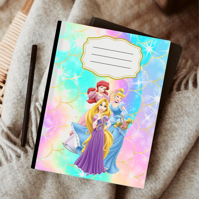 Puede incluir: Un cuaderno de espiral con un dise&ntilde;o colorido que presenta ilustraciones de tres princesas. La portada tiene un fondo pastel con detalles dorados y un &aacute;rea de etiqueta para el nombre del propietario. Un bol&iacute;grafo negro descansa al lado.