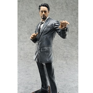 May include: A gray suit figurine of a man with a goatee and glasses. The figurine is posed with one hand raised and the other hand on his hip.