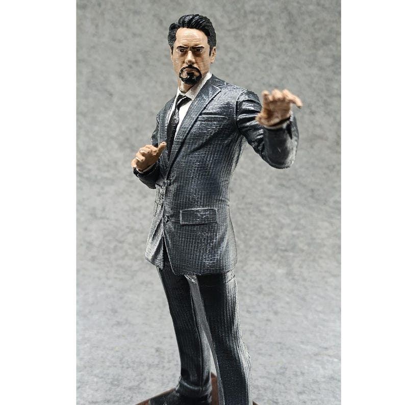 May include: A gray suit figurine of a man with a goatee and glasses. The figurine is posed with one hand raised and the other hand on his hip.