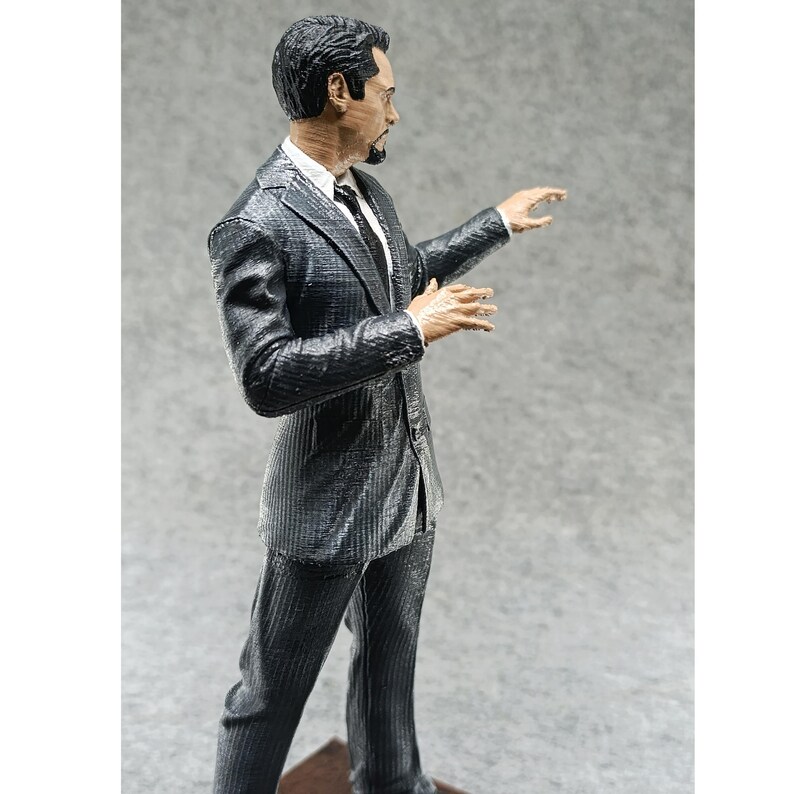 May include: A black and gray suit figure with a black tie. The figure is posed with one arm outstretched and the other hand raised. The figure is made of plastic and is a collectible.