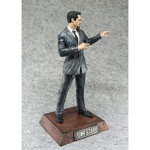 May include: A gray and black figurine of Tony Stark in a suit, standing on a wooden base with the name "TONY STARK" printed on it.
