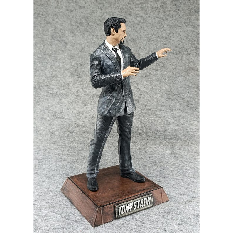 May include: A gray and black figurine of Tony Stark in a suit, standing on a wooden base with the name "TONY STARK" printed on it.