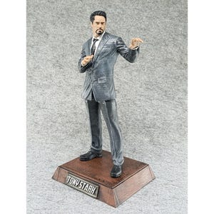 May include: A gray suit figurine of Tony Stark, the Iron Man character, standing on a wooden base with the name "TONY STARK" engraved on it.