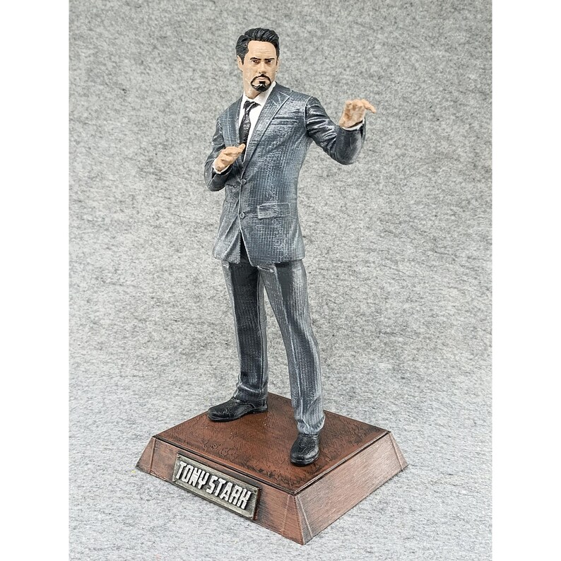 May include: A gray suit figurine of Tony Stark, the Iron Man character, standing on a wooden base with the name "TONY STARK" engraved on it.
