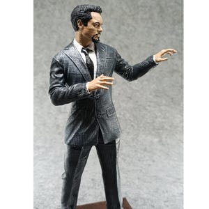 May include: A black and gray suit figurine with a man in a suit standing with his right arm outstretched. The figurine has a detailed face with a beard and a serious expression.