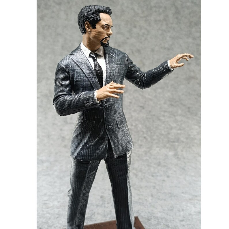 May include: A black and gray suit figurine with a man in a suit standing with his right arm outstretched. The figurine has a detailed face with a beard and a serious expression.