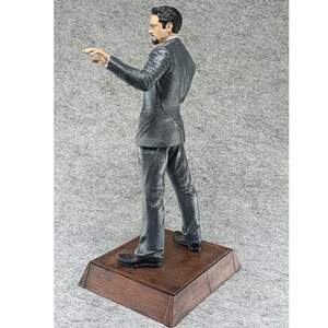May include: A gray suit figurine of a man standing on a wooden base. The man is wearing a dark gray suit with a white shirt and a dark tie. He is looking to the left and has his right hand raised.