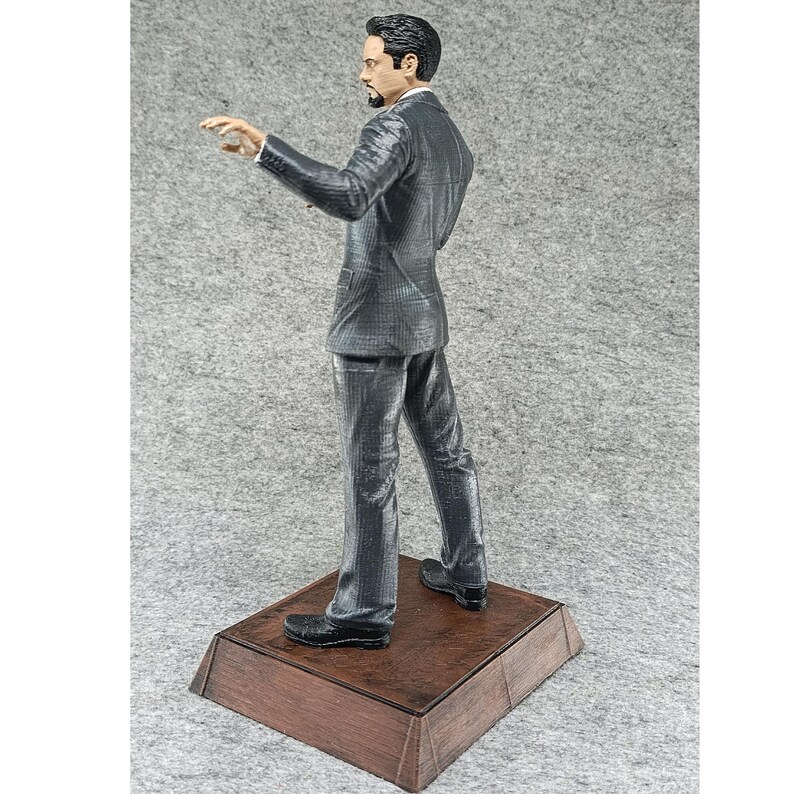 May include: A gray suit figurine of a man standing on a wooden base. The man is wearing a dark gray suit with a white shirt and a dark tie. He is looking to the left and has his right hand raised.