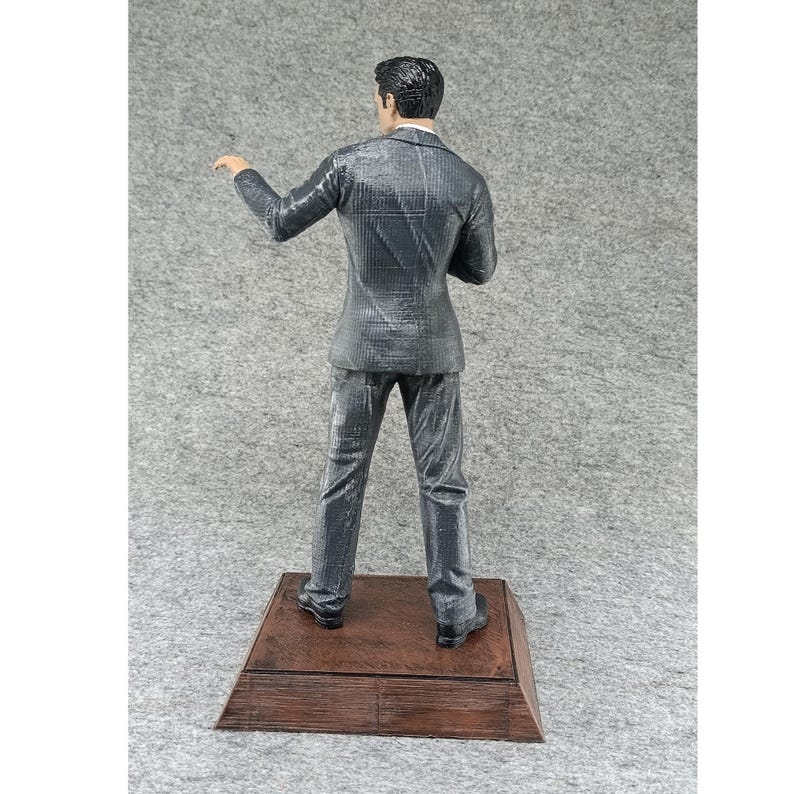 May include: A gray suit figurine standing on a wooden base. The figurine is wearing a gray suit with a pinstripe pattern. The figurine is facing away from the camera.
