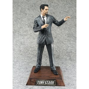 May include: A gray suit figurine of Tony Stark, the Iron Man character, standing on a wooden base with the words "TONY STARK" printed on the front.