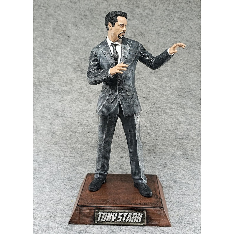May include: A gray suit figurine of Tony Stark, the Iron Man character, standing on a wooden base with the words "TONY STARK" printed on the front.