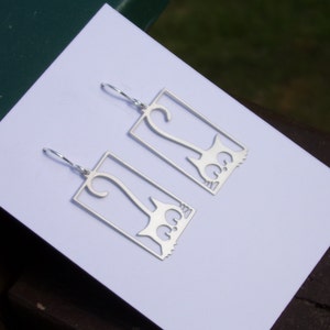 May include: A pair of silver-colored cat-shaped earrings. Each earring features a rectangular frame with a stylized cat silhouette inside. The cat design includes a curled tail and decorative details.
