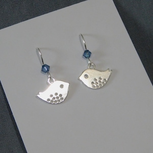 May include: A pair of silver bird-shaped earrings. Each earring has a small, stylized bird pendant with circular indentations. A blue crystal bead hangs above each bird. The earrings are displayed on a white surface.
