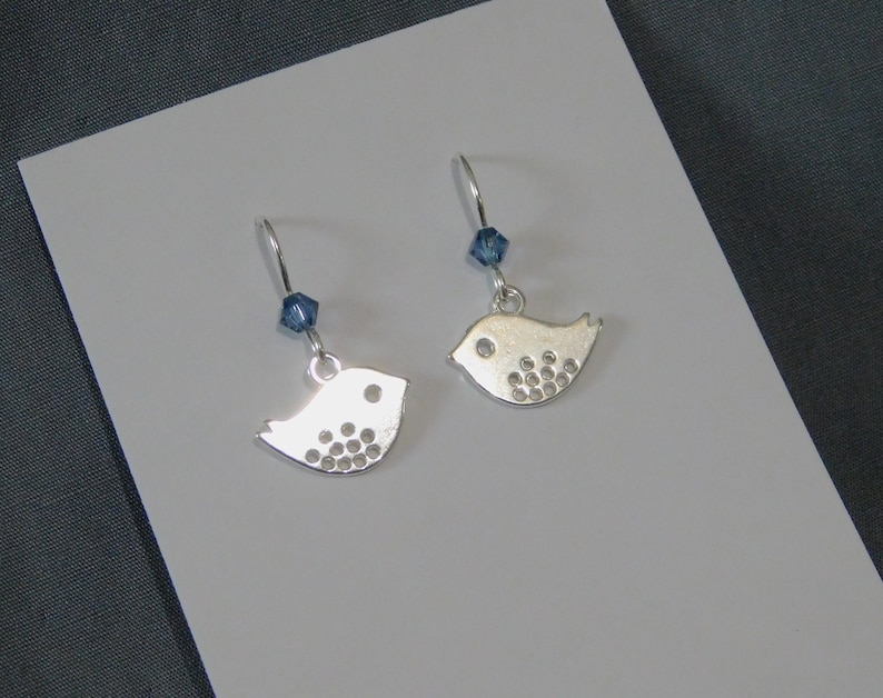 May include: A pair of silver bird-shaped earrings. Each earring has a small, stylized bird pendant with circular indentations. A blue crystal bead hangs above each bird. The earrings are displayed on a white surface.