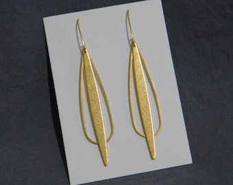 May include: A pair of gold-colored earrings. Each earring features a long, textured, rectangular bar suspended within a teardrop-shaped frame. The earrings are displayed on a white surface against a dark background.