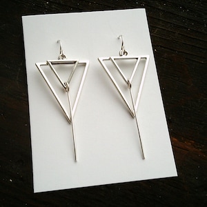May include: A pair of silver geometric earrings. Each earring features a triangular design with a smaller triangle inside and a long, slender bar extending downwards. The earrings are displayed on a white surface.