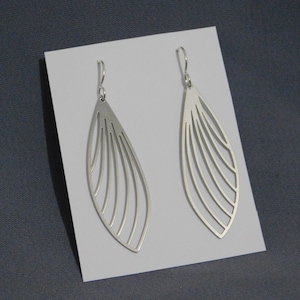 May include: A pair of silver-colored, openwork earrings. Each earring has a long, teardrop shape with a series of curved, parallel lines creating a wing-like design. The earrings are displayed on a white surface against a gray background.