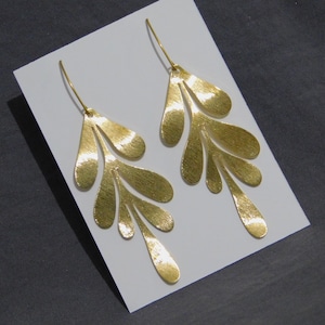 May include: A pair of gold-colored earrings with a leaf-like design. Each earring features multiple textured, elongated teardrop shapes connected to a wire hook. The earrings are displayed on a white surface against a dark gray background.