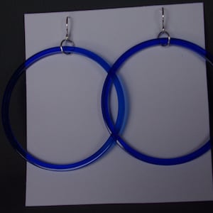 May include: A pair of large, circular blue hoop earrings. The earrings are made of a translucent material and have silver-colored ear wires. The hoops are displayed against a white background.