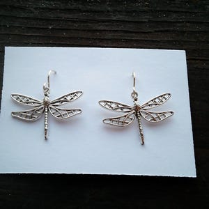May include: A pair of silver dragonfly earrings. Each earring features a detailed dragonfly design with openwork wings and a delicate body. The earrings are displayed on a white surface against a dark wood background.